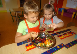 Dwoje dzieci siedzi przy stole. Dziewczynka trzyma w ręku przechylony słoik z miodem, który przelewa chłopcu na łyżkę. Na stole stoi miska z pokrojonymi owocami.